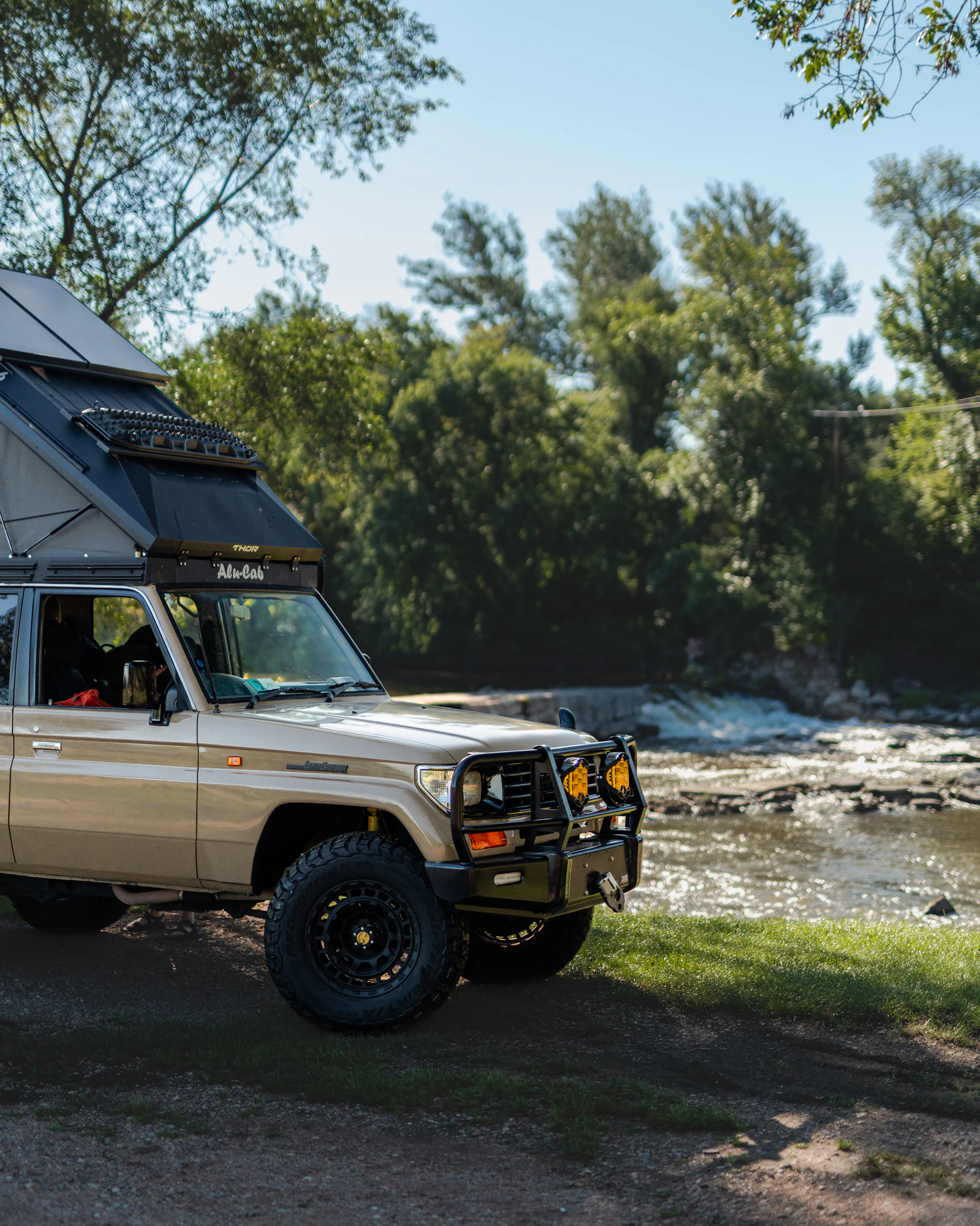 A sand-coloured 4×4 with a rooftop tent parked on a riverbank, trees behind it